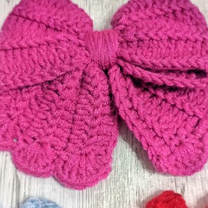Puede incluir: Un lazo de ganchillo grande de color magenta vibrante con una construcci&oacute;n de hilo texturizado y en bucle con un nudo central envuelto. Debajo, se ven parcialmente un lazo de ganchillo azul claro y parte de un lazo rojo brillante.