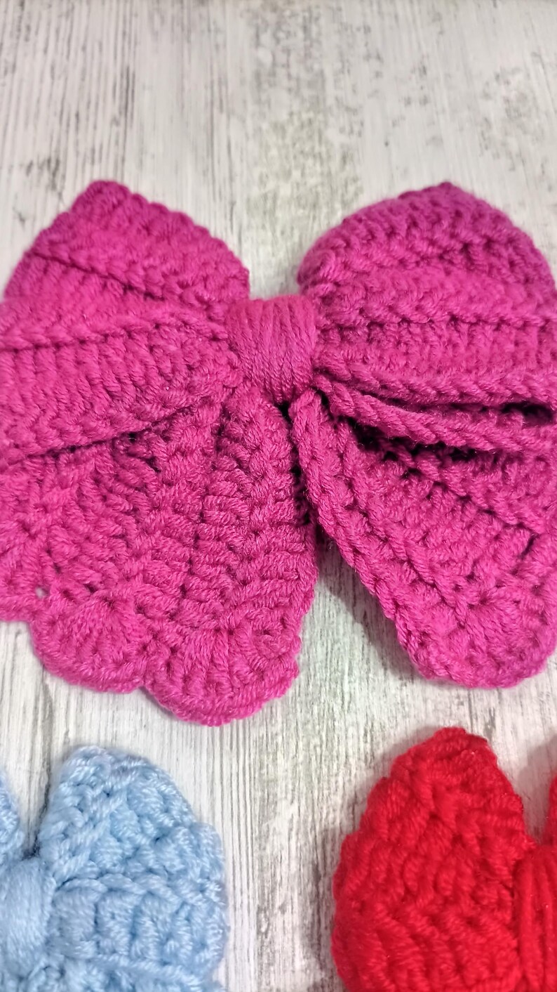 Puede incluir: Un lazo de ganchillo grande de color magenta vibrante con una construcci&oacute;n de hilo texturizado y en bucle con un nudo central envuelto. Debajo, se ven parcialmente un lazo de ganchillo azul claro y parte de un lazo rojo brillante.