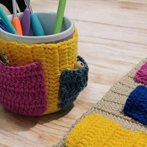 May include: A handmade crochet mug cozy in yellow, with a magenta pocket and a teal accent. A safety pin is attached to the pocket. Crochet hooks are inside the mug. A matching crochet coaster with colorful squares is also present.