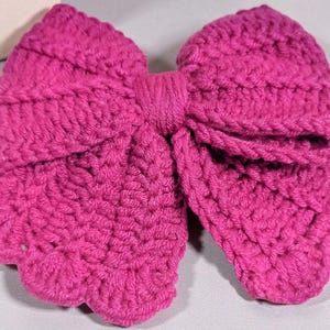 Puede incluir: Un lazo de ganchillo en color magenta vibrante con una textura de hilo tejido a mano. El accesorio presenta dos grandes bucles redondeados y un nudo central, mostrando un patr&oacute;n de puntadas denso y uniforme en toda su estructura.