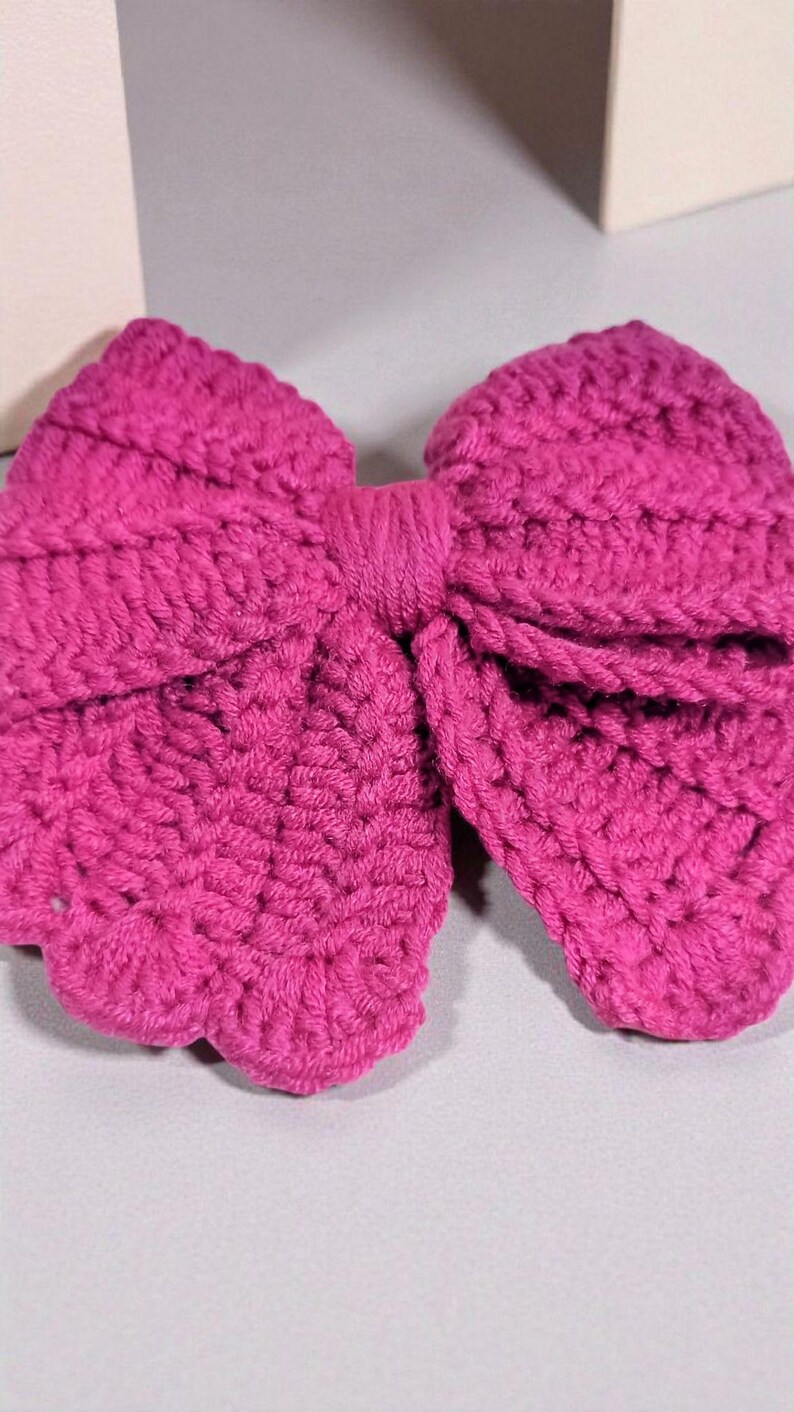 Puede incluir: Un lazo de ganchillo en color magenta vibrante con una textura de hilo tejido a mano. El accesorio presenta dos grandes bucles redondeados y un nudo central, mostrando un patr&oacute;n de puntadas denso y uniforme en toda su estructura.