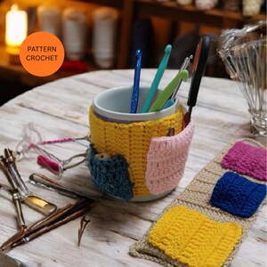 May include: A white mug with a yellow crocheted cozy and three small pockets in blue, pink, and yellow. The mug holds crochet hooks and a pair of scissors. The text "PATTERN CROCHET" is in an orange circle.