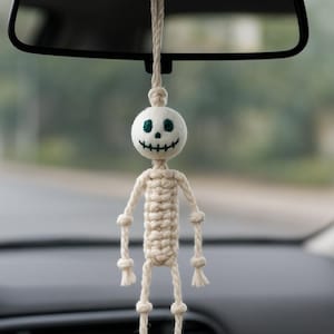 May include: A macramé skeleton car charm suspended from a car's rearview mirror. The skeleton features a white head with green eyes and a stitched smile. The body and limbs are made of knotted beige rope.