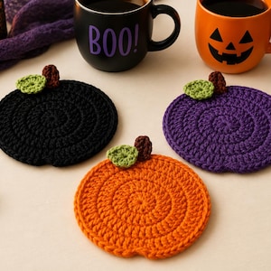 Peut inclure: Trois dessous de verre en crochet en forme de citrouilles, de couleurs noire, violette et orange. Chaque dessous de verre présente une petite feuille verte et une tige marron. Ils sont disposés près d'un mug noir avec le texte BOO! et un mug orange.