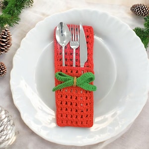 May include: A red crocheted cutlery holder with a green bow, holding a spoon, fork, and knife. The holder is placed on a white plate, with pine cones and greenery in the background. A festive table setting.