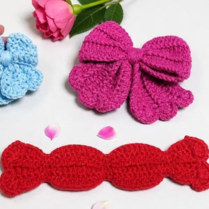Puede incluir: Un accesorio de ganchillo azul claro en forma de flor, un gran lazo de ganchillo magenta y una pieza larga de ganchillo roja con borde festoneado. Estos tres art&iacute;culos textiles distintos est&aacute;n hechos con puntos visibles.