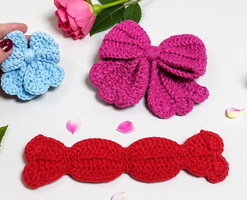 Puede incluir: Un accesorio de ganchillo azul claro en forma de flor, un gran lazo de ganchillo magenta y una pieza larga de ganchillo roja con borde festoneado. Estos tres art&iacute;culos textiles distintos est&aacute;n hechos con puntos visibles.