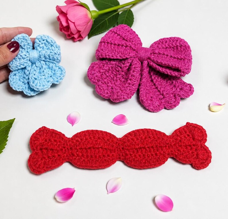 Puede incluir: Una flor de ganchillo azul claro, un lazo de ganchillo magenta grande y una tira de ganchillo roja larga con extremos redondeados. Los art&iacute;culos est&aacute;n dispuestos con una rosa y p&eacute;talos rosas esparcidos, mostrando texturas de hilo.