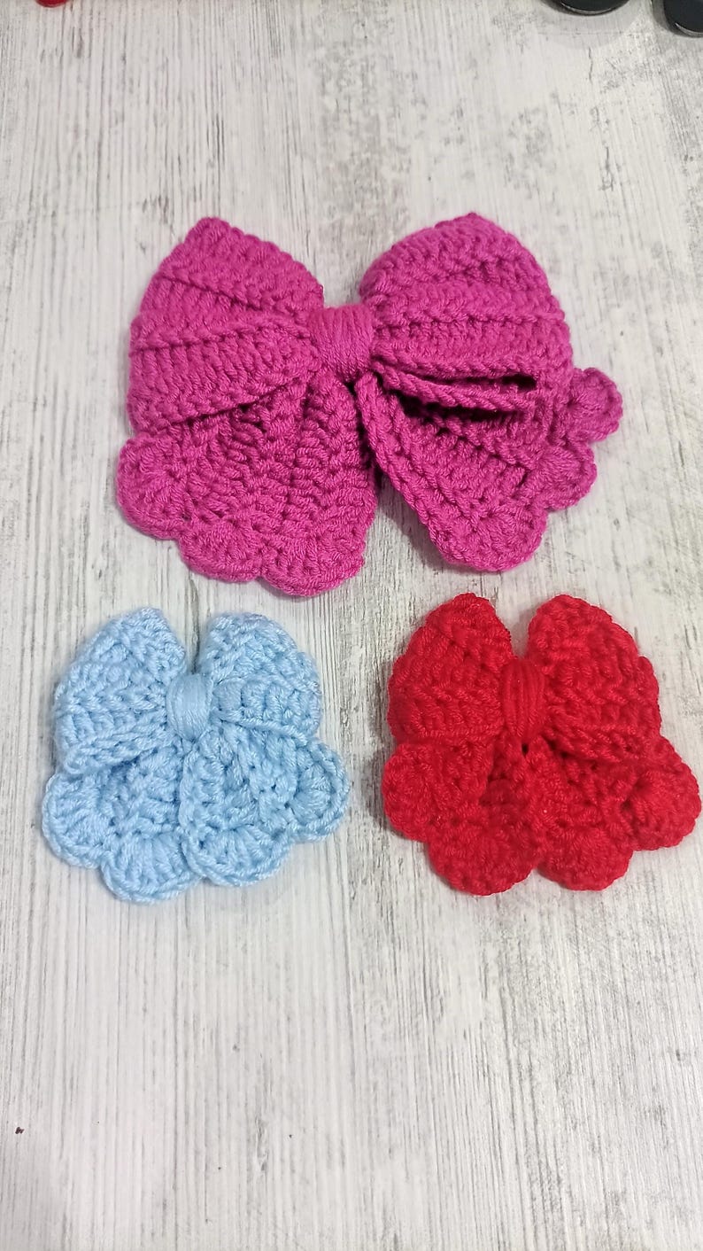 Puede incluir: Tres lazos de ganchillo hechos a mano en rosa brillante, azul claro y rojo. El lazo rosa grande est&aacute; colocado en la parte superior, mientras que los lazos m&aacute;s peque&ntilde;os azul claro y rojo est&aacute;n dispuestos uno al lado del otro debajo. Borde festoneado.