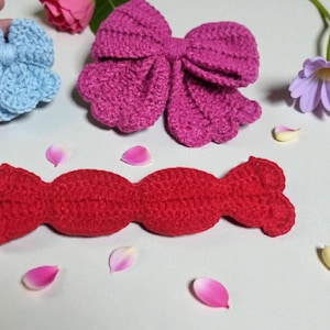 Puede incluir: Una flor de ganchillo azul claro, un lazo de ganchillo magenta grande y una pieza de ganchillo roja larga con forma de caramelo. Estos tres art&iacute;culos textiles hechos a mano est&aacute;n dispuestos con p&eacute;talos de flores y una hoja verde.