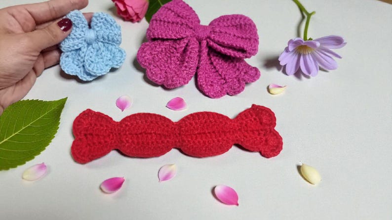 Puede incluir: Una flor de ganchillo azul claro, un lazo de ganchillo magenta grande y una pieza de ganchillo roja larga con forma de caramelo. Estos tres art&iacute;culos textiles hechos a mano est&aacute;n dispuestos con p&eacute;talos de flores y una hoja verde.