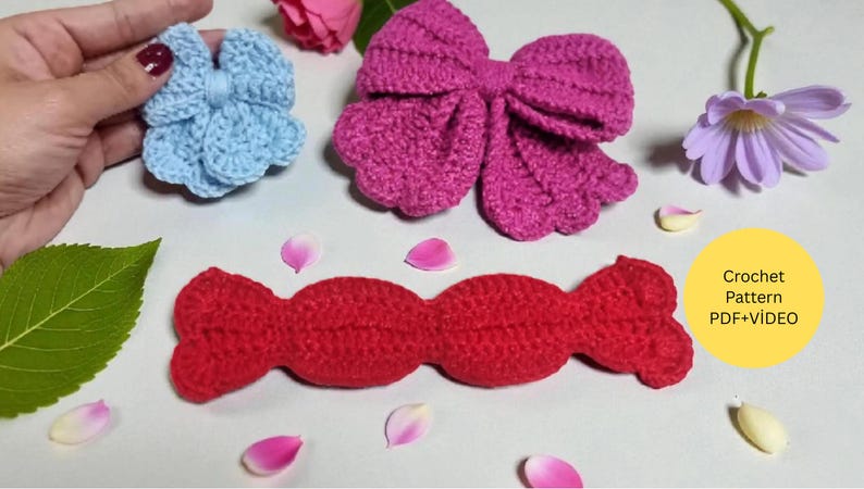 Puede incluir: Un lazo de ganchillo azul claro, un lazo de ganchillo magenta grande y una pieza de ganchillo roja larga con forma de caramelo. Un c&iacute;rculo amarillo contiene el texto Crochet Pattern PDF+VIDEO. Los objetos est&aacute;n dispuestos con una hoja verde y p&eacute;talos.