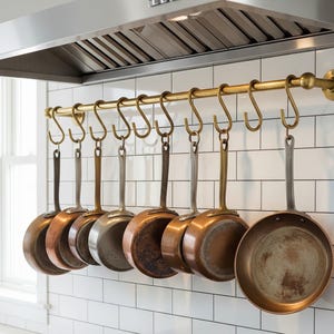 May include: A collection of copper and stainless steel pots and pans hanging from a brass rod with S-hooks. The kitchenware is suspended against a white tiled wall, beneath a stainless steel range hood.