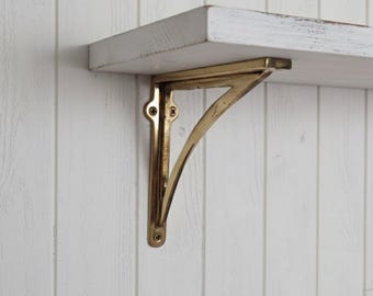 May include: A gold-colored metal shelf bracket is mounted on a white wooden wall. The bracket is a decorative support for a shelf, with a curved design and visible screw heads. The shelf is white with a distressed finish.