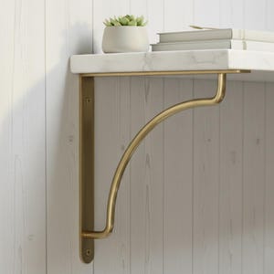 May include: A gold-colored metal shelf bracket with a curved support. The bracket is mounted on a white wooden wall and supports a white marble shelf with a small potted plant and books.