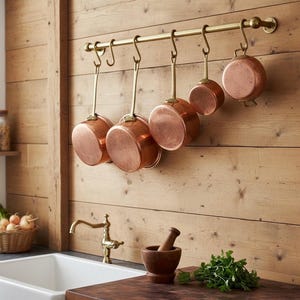 May include: A collection of copper pots hanging from a brass rod on a wooden wall. The pots vary in size and shape, with shiny copper exteriors and brass handles. The kitchen setting includes a sink, faucet, and a mortar and pestle.