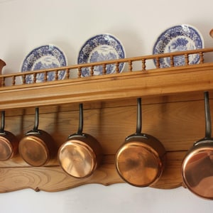 Op de afbeelding: Een houten pottenrek met koperen pannen onder een plank met blauw-witte borden en bruine keramische kruiken. Het rek is aan een witte muur bevestigd, wat een rustieke keukenesthetiek creëert.