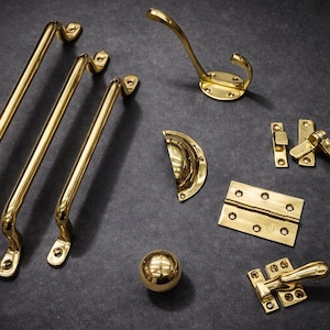 Könnte beinhalten: Eine Sammlung von polierten Messingbeschlägen, darunter Türgriffe, ein Kleiderhaken, ein Schrankgriff, Scharniere, ein Türgriff und ein kugelförmiger Knopf. Die Beschläge sind auf einer dunkelgrauen Oberfläche angeordnet.