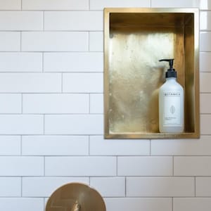 Puede incluir: Una botella blanca con un dispensador de bomba negro se encuentra dentro de un estante empotrado de color dorado. La botella tiene la palabra "Botanica" impresa, junto con un gráfico de hoja. El estante está colocado en una pared de azulejos blancos.