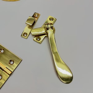 A collection of polished brass hardware components, including a handle, hinges, and a pull، The pieces have a bright, reflective finish and are designed for furniture or cabinetry. The hardware is new and unused.