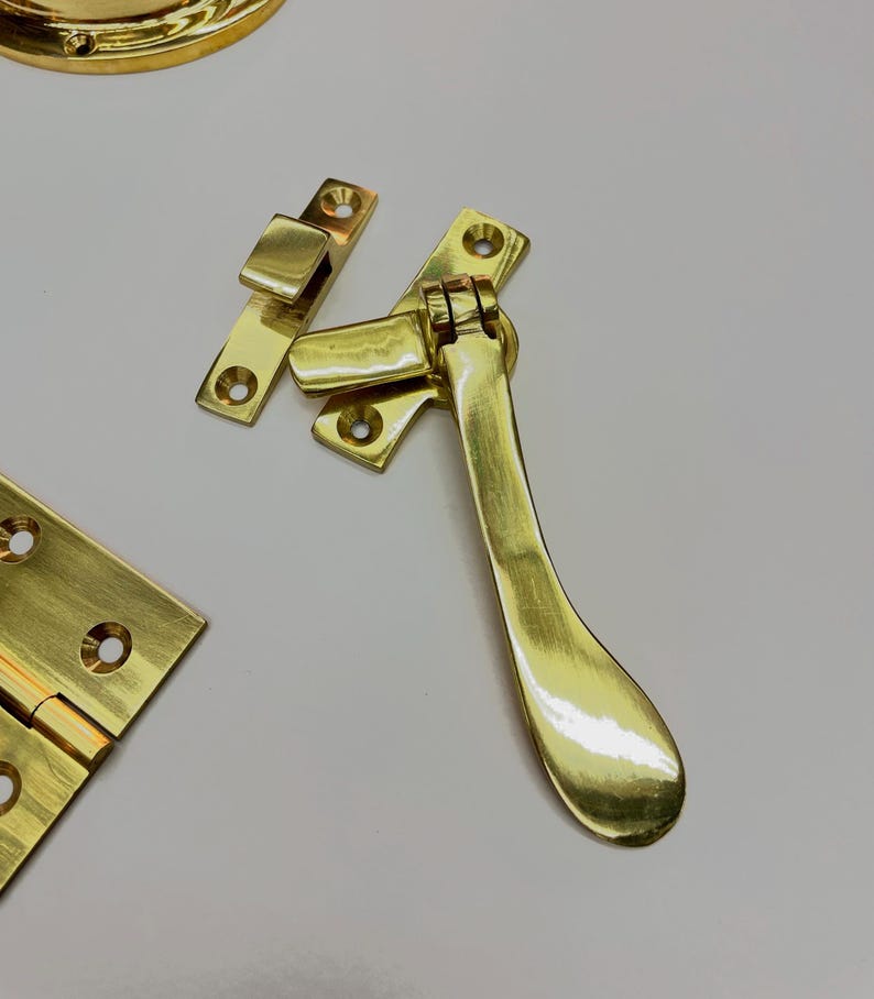 A collection of polished brass hardware components, including a handle, hinges, and a pull، The pieces have a bright, reflective finish and are designed for furniture or cabinetry. The hardware is new and unused.