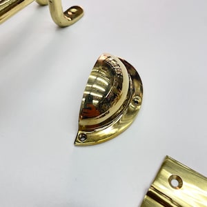 Close-up of several brass hardware pieces. including a half-moon pull, a cabinet handle, and a hinge. The hardware has a shiny, reflective surface. The items are arranged on a white surface.