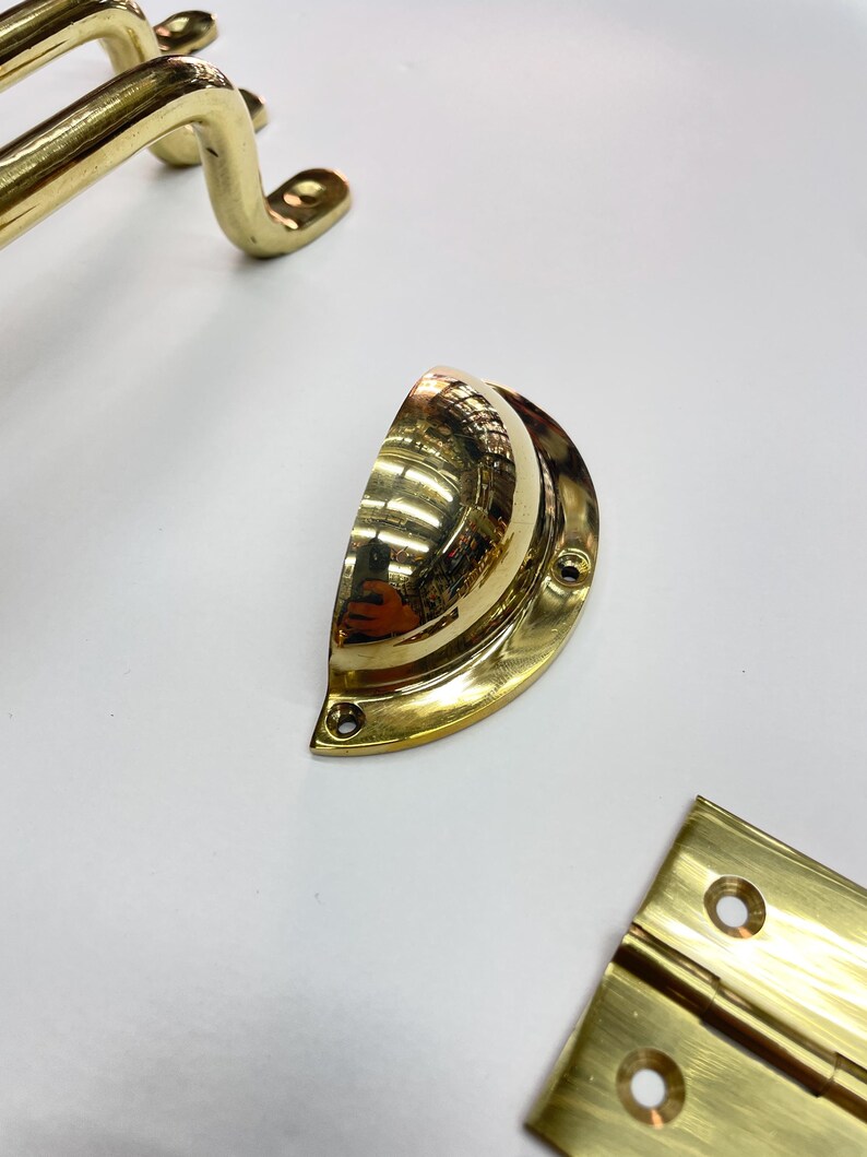 Close-up of several brass hardware pieces. including a half-moon pull, a cabinet handle, and a hinge. The hardware has a shiny, reflective surface. The items are arranged on a white surface.
