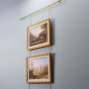 Könnte beinhalten: Zwei gerahmte Landschaftsbilder mit goldenen, verzierten Rahmen hängen über Ketten an einer goldenen Stange. Die Gemälde zeigen Berg- und Küstenszenen. Die Wand ist in einem gedämpften Blau-Grau gestrichen. Die goldene Stange ist horizontal montiert.