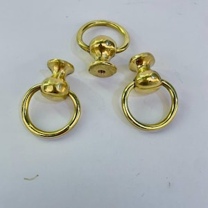 May include: Three brass-colored ring pulls. Each pull features a rounded base with a ring attached. The pulls are made of a shiny, metallic material. The ring pulls are likely used for furniture or decorative purposes.