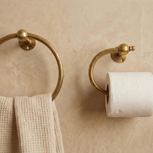 Handmade solid brass towel ring and toilet paper holder set with a polished gold finish, mounted on a neutral tadelakt wall