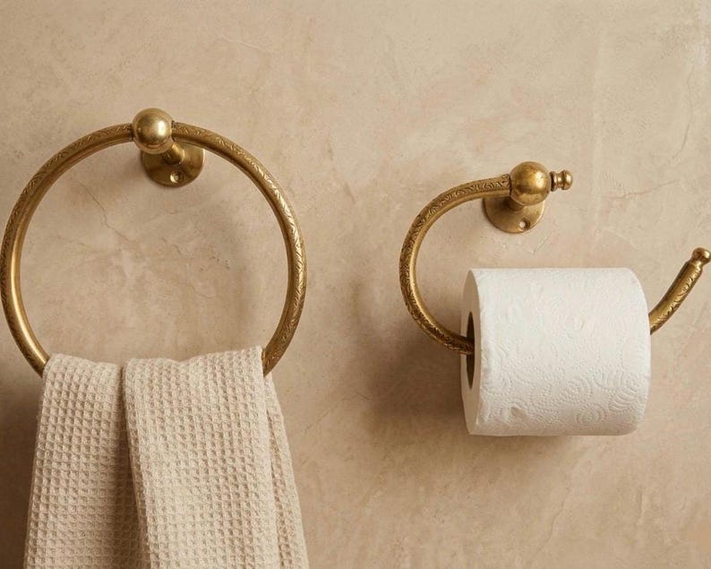 Handmade solid brass towel ring and toilet paper holder set with a polished gold finish, mounted on a neutral tadelakt wall