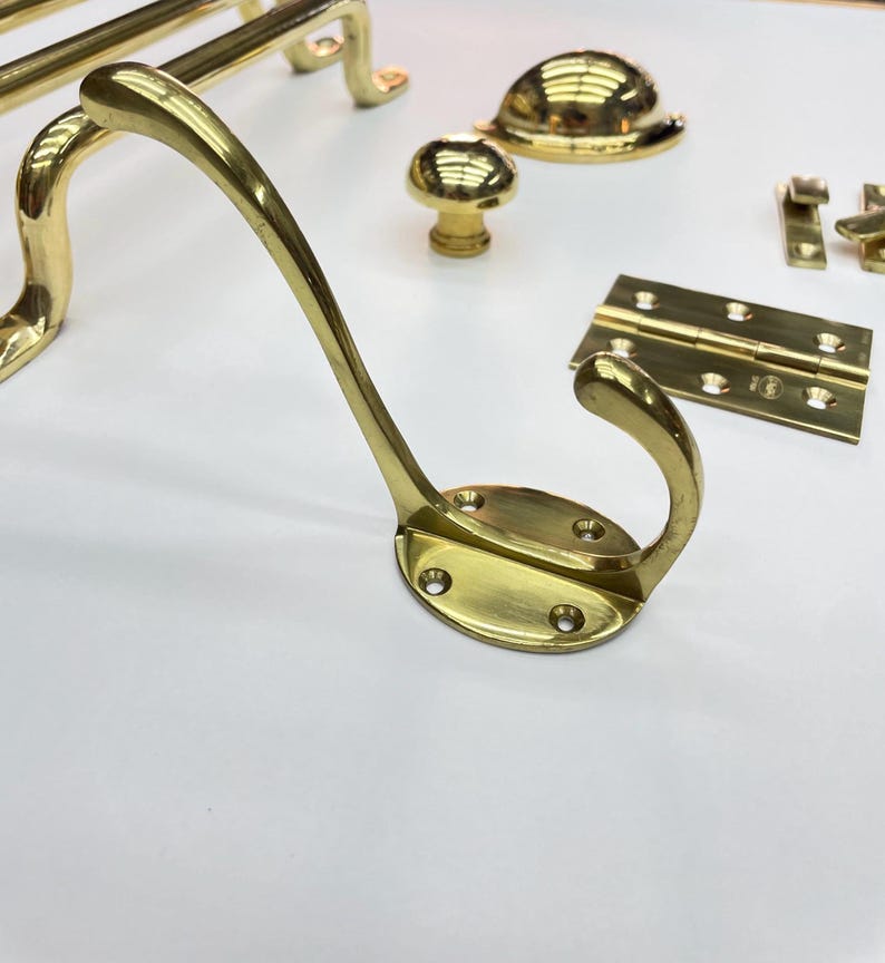 A collection of polished brass hardware, including a coat hook, cabinet knobs،door handles, and hinges.The hardware is arranged on a white surface, showcasing the reflective quality of the brass.