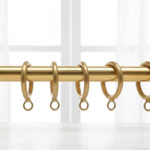 以下が含まれることがあります： 金色のカーテンレールと、それに合うリング。 レールは水平で、リングは等間隔に配置されています。 リングは、カーテンを取り付けるための小さなループが付いた、シンプルで丸みを帯びたデザインです。 背景は明るくぼやけた窓です。