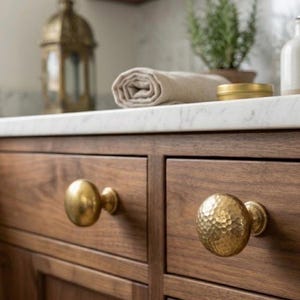 May include: Close-up of a wooden cabinet with two drawers and brass knobs. The cabinet is made of dark wood and has a marble countertop. A rolled towel, small plant, and decorative items are on the countertop.