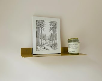 May include: A gold-colored metal shelf mounted on a pink wall, holding a framed black and white landscape print and a glass jar candle with a gold lid. The candle has text on the label.