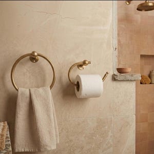 Handmade solid brass towel ring and toilet paper holder set with a polished gold finish, mounted on a neutral tadelakt wall