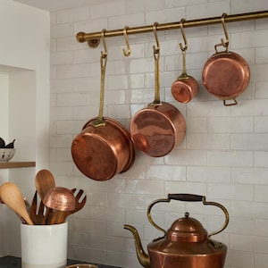 May include: Copper kitchenware set hanging on a brass rail against a white tiled wall. The set includes pots, a small saucepan, and a kettle. Wooden utensils are in a white ceramic container.