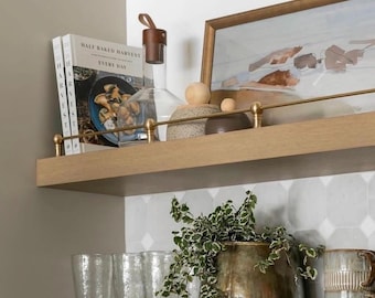 May include: Two wooden shelves displaying home decor. The top shelf features a glass bottle with a brown stopper, a framed painting, and books. The lower shelf holds glasses, a plant in a bronze pot, and mugs.
