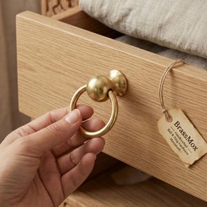 Könnte beinhalten: Nahaufnahme eines Messing-Schubladengriffs mit Ringgriff. Der Griff ist an einer hellen Holzschublade befestigt. Ein Etikett mit dem Text "BrassMox" hängt an der Schublade. Die Schublade ist teilweise geöffnet und zeigt gefalteten Stoff.