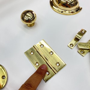 Może przedstawiać: Zbi&oacute;r błyszczących, złotych element&oacute;w okuć, w tym gałka do szafki, uchwyt, zawias i uchwyt wpuszczany. Okucia są ułożone na białej powierzchni, prezentując ich odblaskowe powierzchnie i r&oacute;żne kształty.