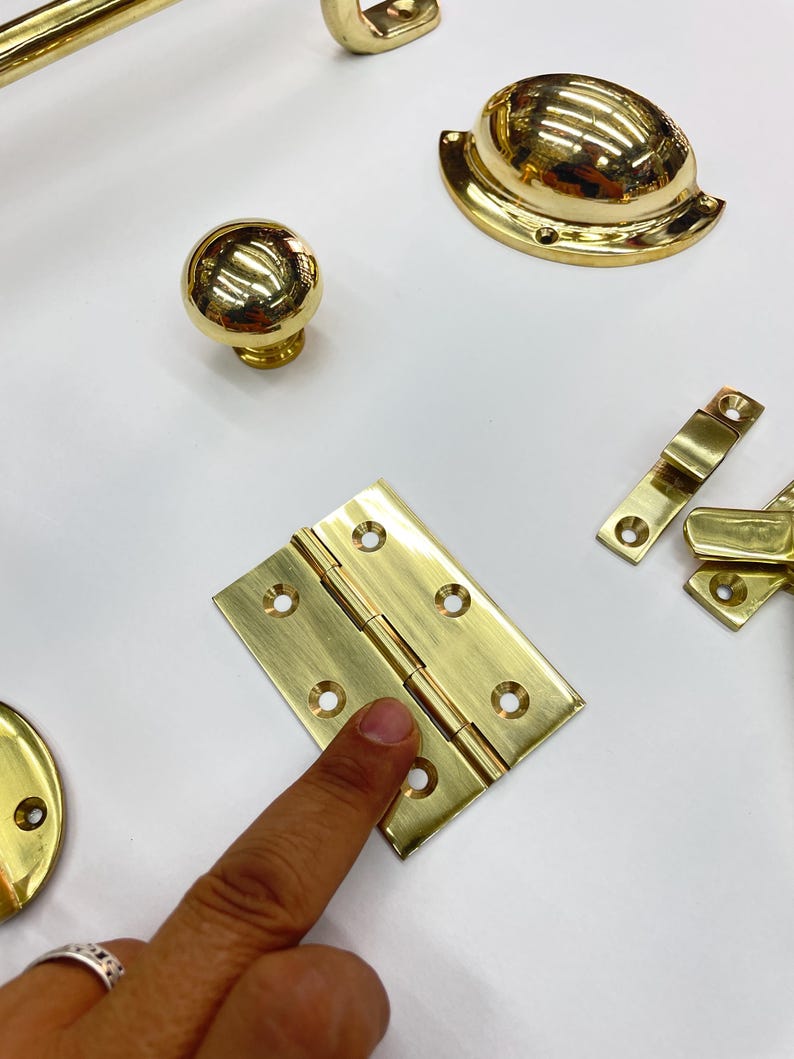 Może przedstawiać: Zbi&oacute;r błyszczących, złotych element&oacute;w okuć, w tym gałka do szafki, uchwyt, zawias i uchwyt wpuszczany. Okucia są ułożone na białej powierzchni, prezentując ich odblaskowe powierzchnie i r&oacute;żne kształty.