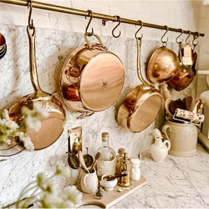 Peut inclure: Des ustensiles de cuisine en cuivre, dont des casseroles et des poêles, sont suspendus à une barre en laiton contre un mur à motifs de marbre. Divers accessoires de cuisine, tels que des bouteilles et de petits récipients, sont disposés sur une planche à découper en bois.