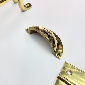 Close-up of several brass hardware pieces, including a curved handle, a hinge, and other curved pieces. The hardware has a shiny. reflective surface and appears to be new. The pieces are arranged on a white surface