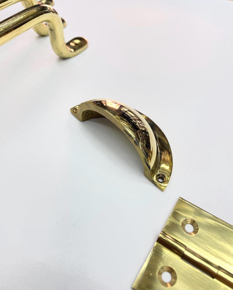 Close-up of several brass hardware pieces, including a curved handle, a hinge, and other curved pieces. The hardware has a shiny. reflective surface and appears to be new. The pieces are arranged on a white surface