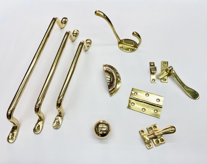 Może przedstawiać: Kolekcja polerowanych element&oacute;w okuć mosiężnych, w tym uchwyt&oacute;w do drzwi, wieszaka na płaszcze, gałki do szafki, zawias&oacute;w i okuć do okien. Okucia mają jasne, odblaskowe wykończenie i są ułożone na białej powierzchni.