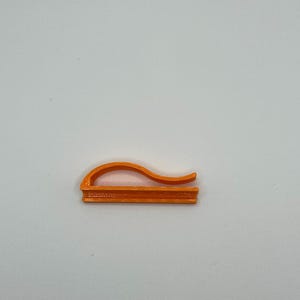 May include: An orange, 3D-printed clip with a curved top and a flat base. The clip is designed to hold items together. The object is set against a plain white background.