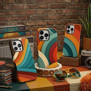 May include: Three smartphone cases featuring a layered, curved graphic pattern in shades of orange, teal, cream, and dark red. The cases have a cut-out for the camera lens and are displayed alongside a vintage radio, a camera, and sunglasses.