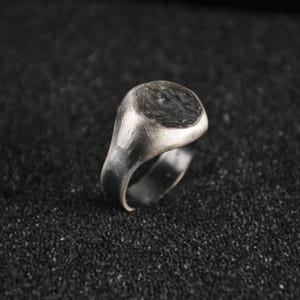 Könnte beinhalten: Ein silberner Siegelring mit einer dunklen, strukturierten ovalen Oberfläche. Der Ring hat ein gebürstetes Metall-Finish und ein klassisches, minimalistisches Design. Der Ring ist vor einem dunklen, strukturierten Hintergrund platziert.