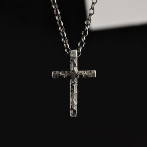 May include: A silver cross pendant with a textured finish, suspended from a matching chain. The cross is set against a dark backdrop, emphasising its detailed design. The necklace is a religious item.