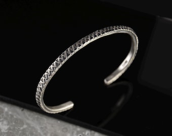 May include: A silver bracelet with a textured, geometric pattern. The bracelet is open and has a simple, minimalist design.